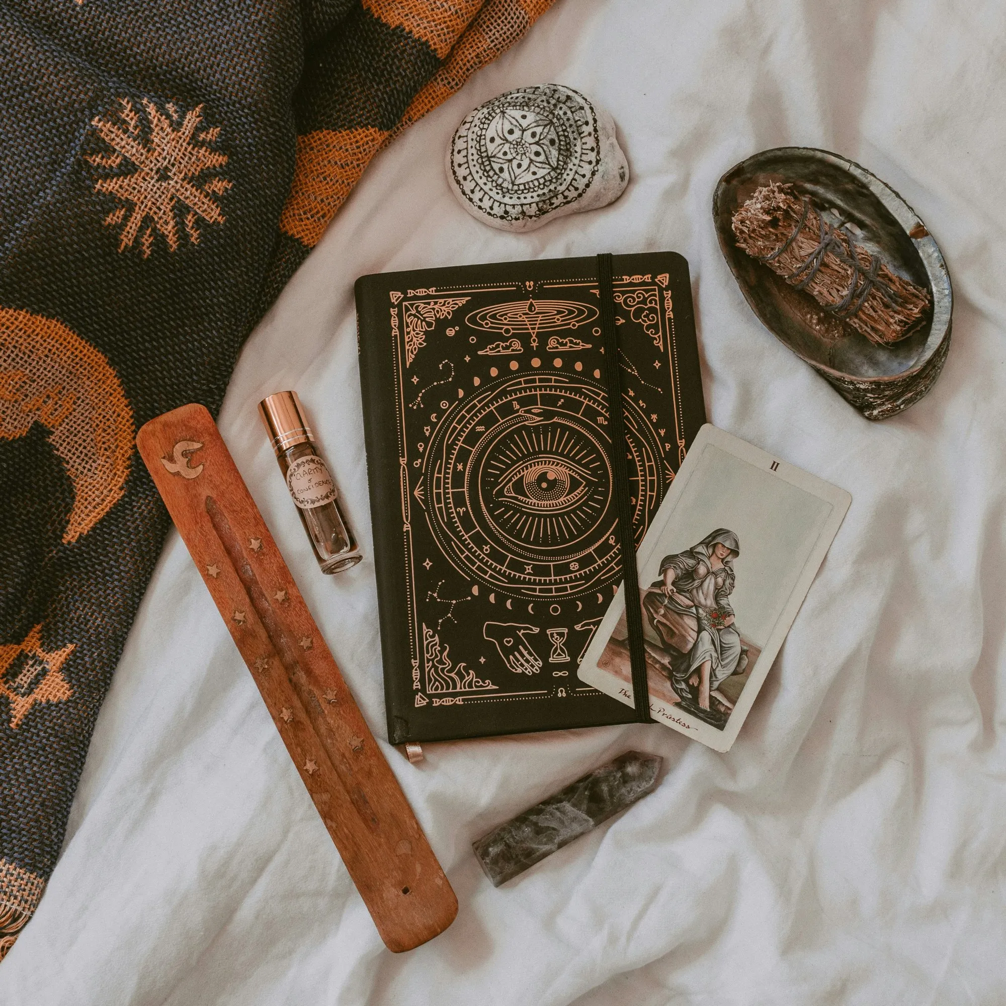 Carnet à l’œil ésotérique, carte de tarot, sauge, pierre peinte et porte-encens posés sur un drap blanc